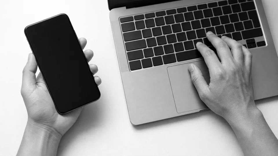 A hand holding a smartphone and a hand on a laptop keyboard